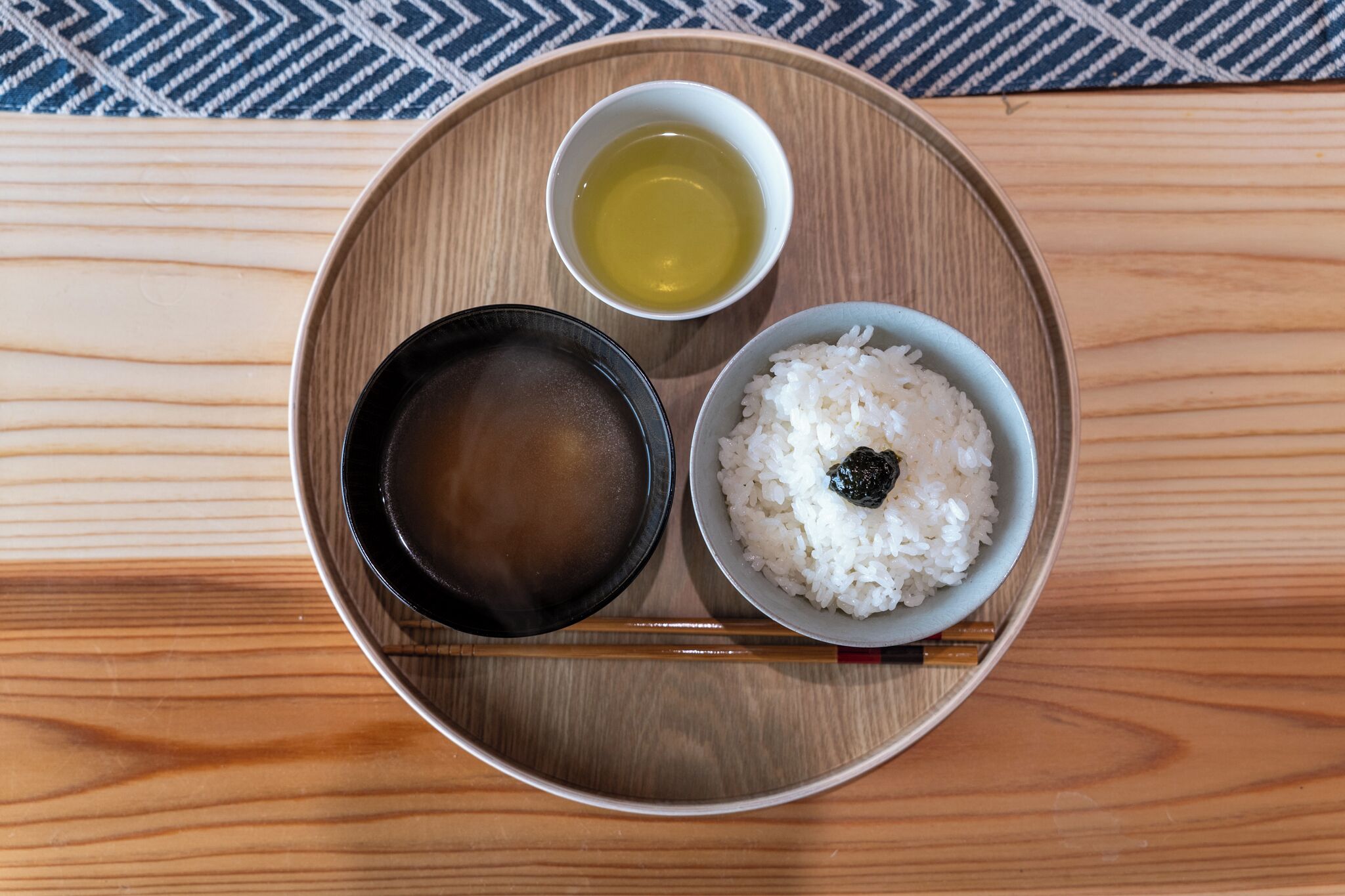 A wooden tray with a bowl of white rice, a small bowl of soy sauce, and a cup of tea on a wooden table.