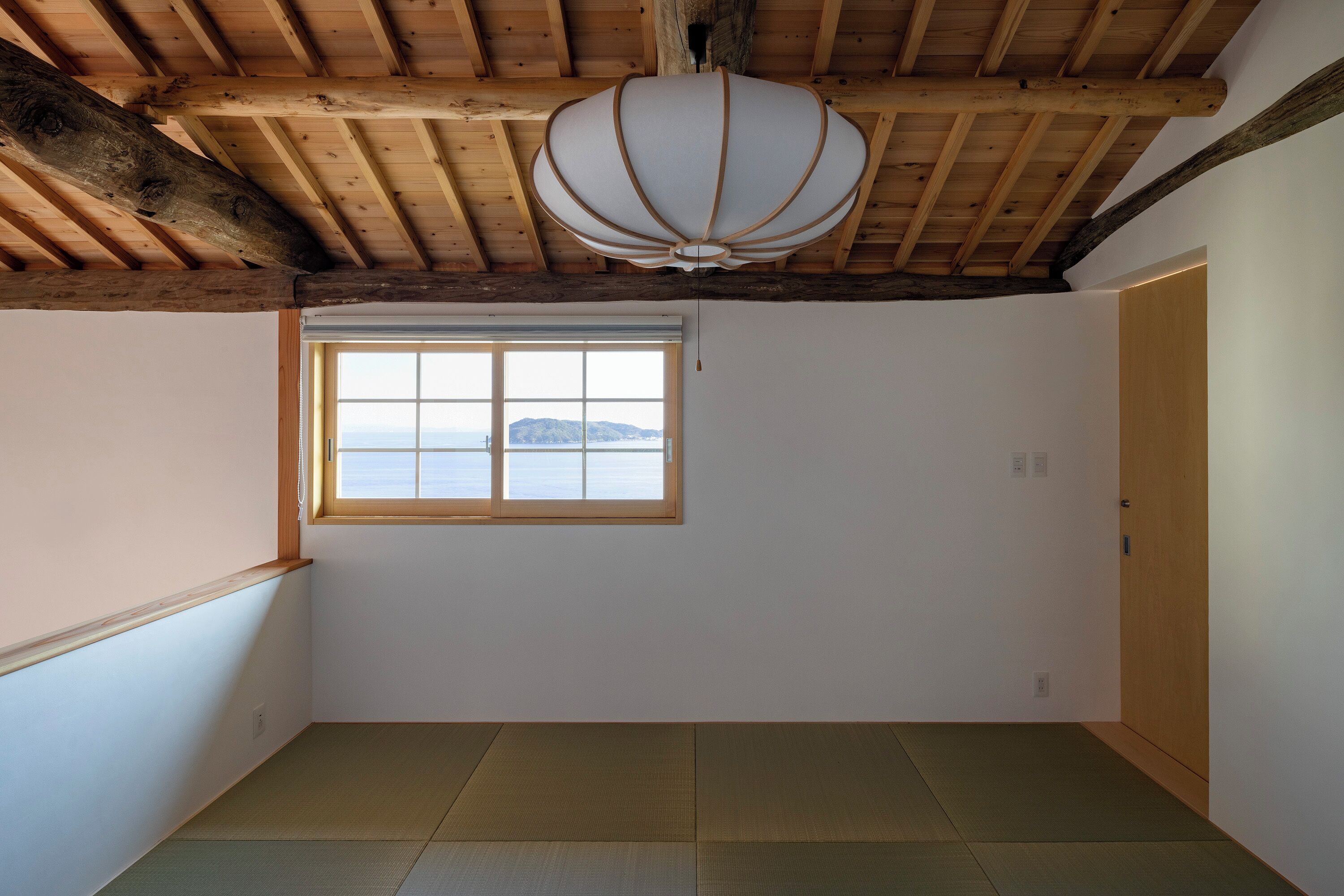 Una habitación con suelo de tatami, techo de madera, ventana con vistas y un farol japonés tradicional de papel.