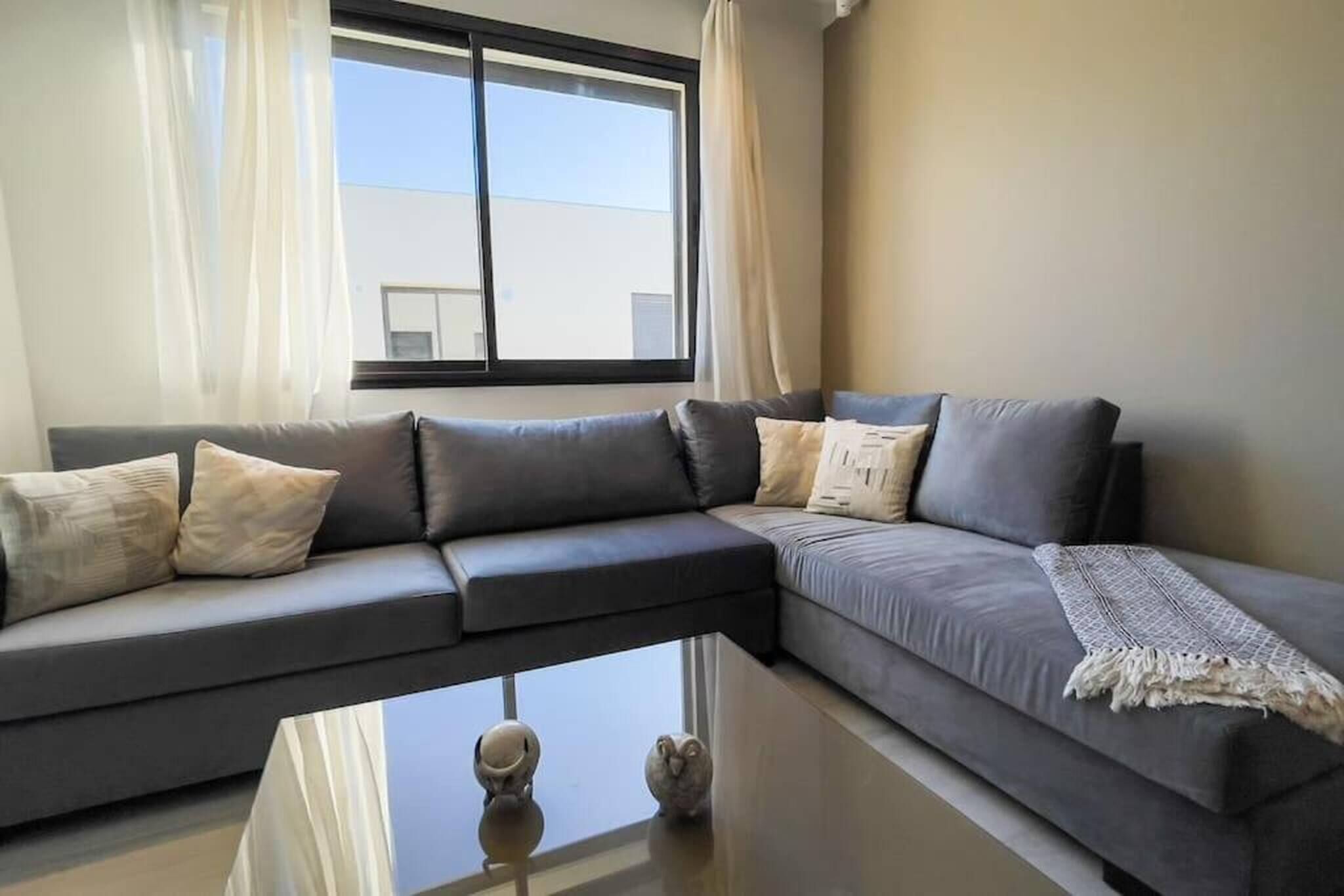 A modern living room with a grey sectional sofa, a glass coffee table, and a view of a building through a large window.