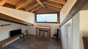 A cozy room with a sloped ceiling, a window with a mountain view, a wooden table with two chairs, a TV on a stand, and a bench. - Enchantment of the Lake app Palanzo Studio Apartment with Terrace A/C (Molina)