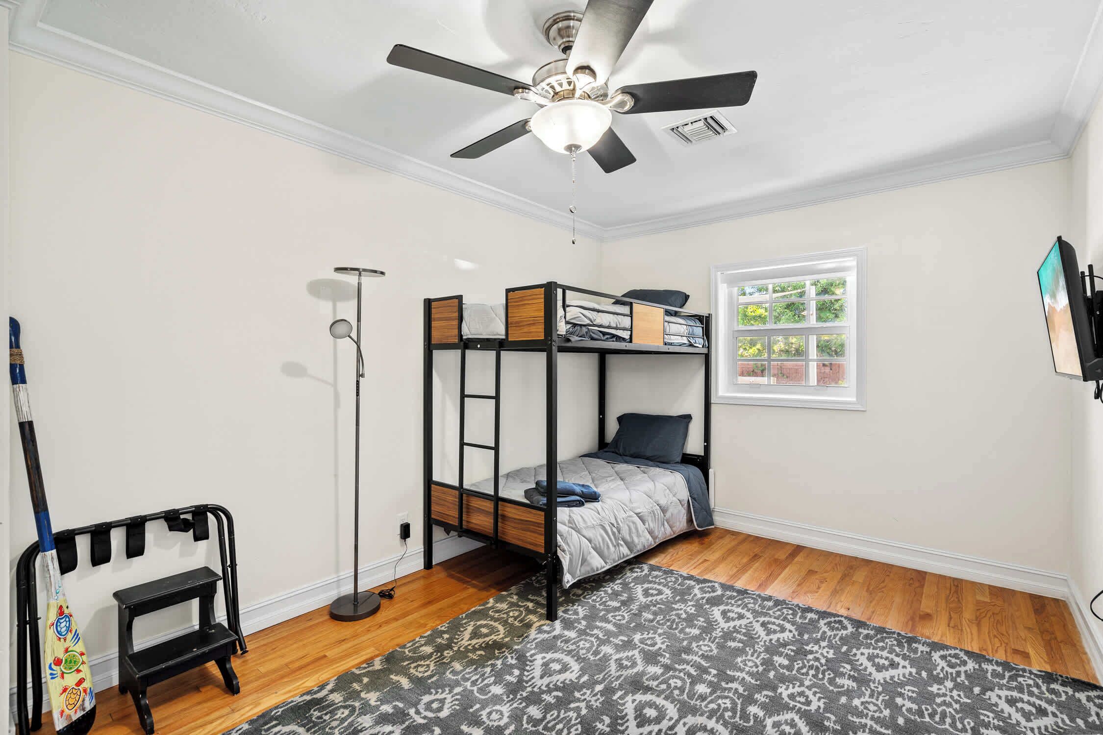 A bedroom with a bunk bed, a standing fan, a window with a view, a patterned rug, and a wall-mounted TV.
