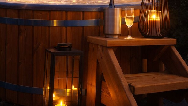Un bain à remous avec une bougie et un récipient en métal, entouré de structures en bois et illuminé de guirlandes lumineuses.