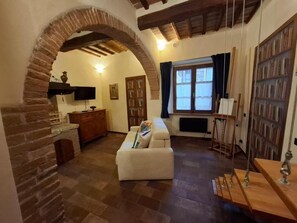 A cozy living room with a fireplace, a sofa, a bookshelf, and a window with curtains. - Il Pacchiarotto-Medieval Walls (Casole d'Elsa)