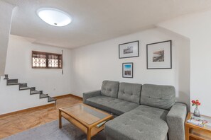 Ein Wohnzimmer mit grauem Sofa, einem Glastisch, einer Treppe und einem Fenster mit Holzgitter.