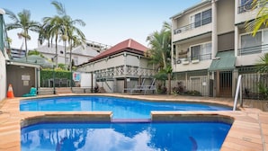 Uma piscina com deck revestido, um edifício com telhado vermelho e palmeiras.