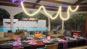 Un espace de restauration en plein air avec des tables dressées pour un repas, une piscine en arrière-plan et des guirlandes lumineuses suspendues au-dessus.