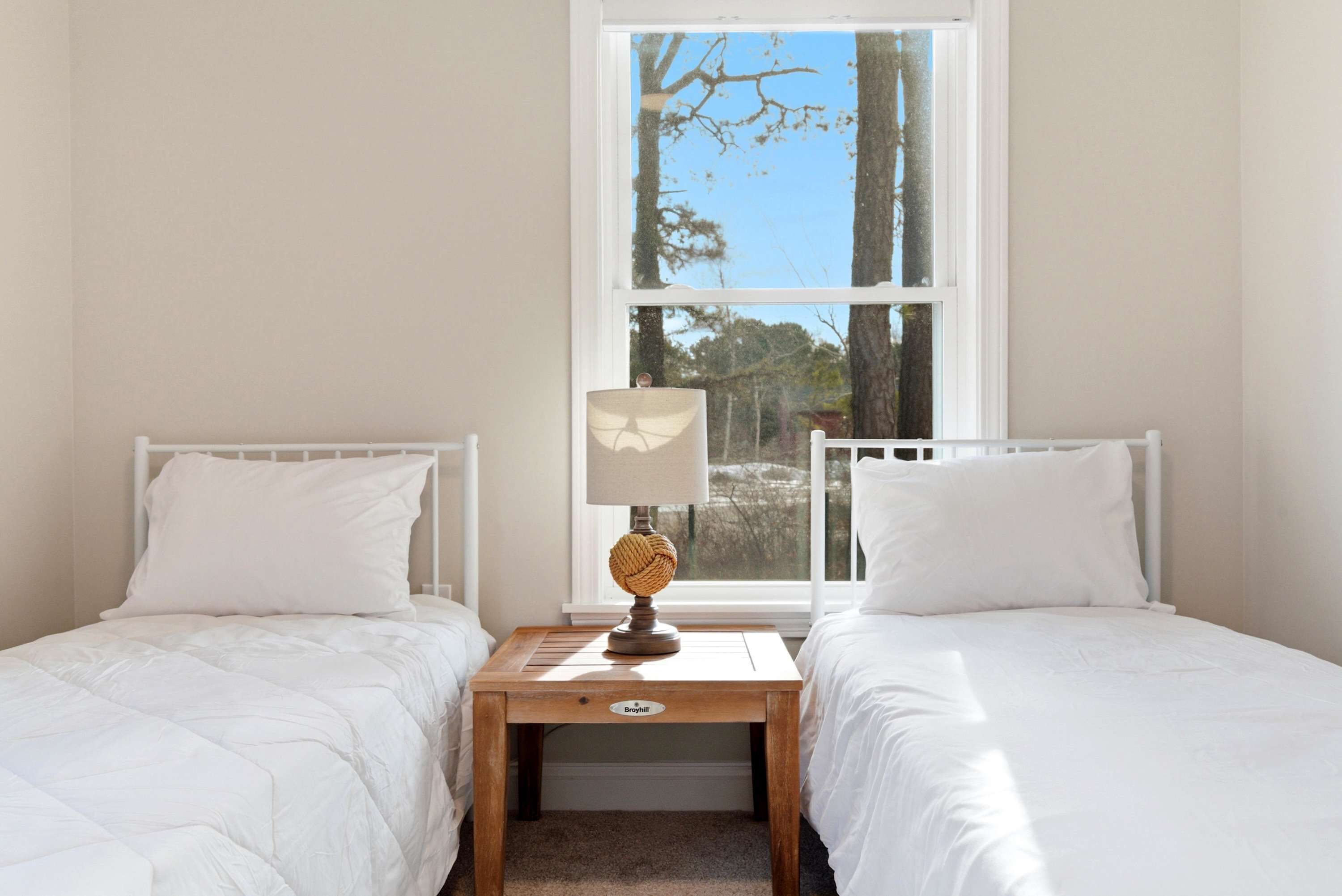 A bedroom with two beds, a nightstand with a lamp, and a window with a view of trees.
