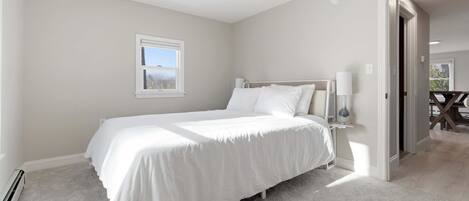 A bedroom with a single bed, a bedside table with a lamp, a window, and a door leading to another room.
