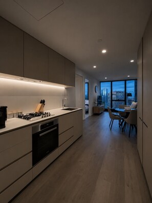 A modern kitchen with a gas stove, oven, and sink, leading to a dining area with a view of a cityscape. - Skyline Suites | Floor-to-Ceiling Views, Level 38 (Southbank)