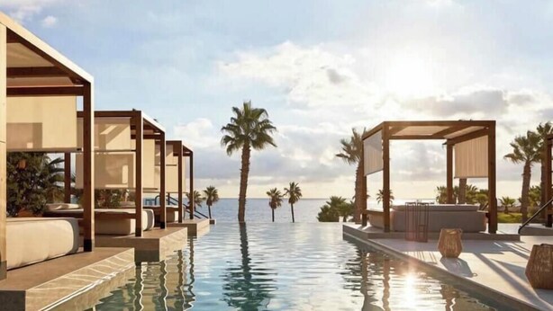 Un área junto a la piscina con sillones de descanso y sombrillas, palmeras y un cielo despejado.