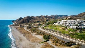 Un resort costero con acceso a la playa, una carretera que lleva a un conjunto de edificios blancos, y montañas al fondo.