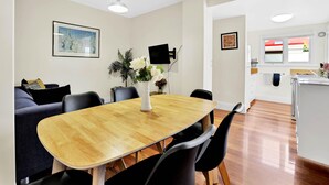 A dining area with a wooden table and chairs, a sofa, a wall-mounted TV, and a kitchen in the background.