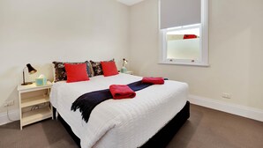 A neatly made bed with a white comforter and red accents, a bedside table with a lamp, and a window with a view.