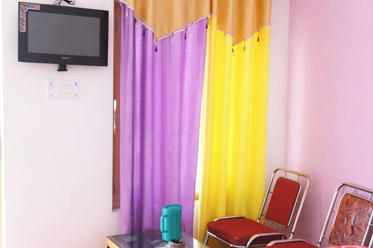 Une chambre avec un lit, un téléviseur fixé au mur, une petite table avec une chaise et des rideaux colorés.