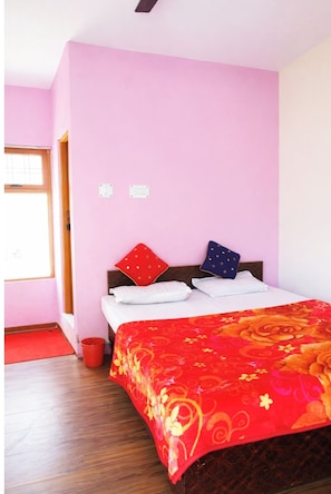 A bedroom with a bed, pillows, a red throw blanket, a wooden floor, a red bucket, and a window. - Hotel Utsav Palace Uttarkashi (Dunda)