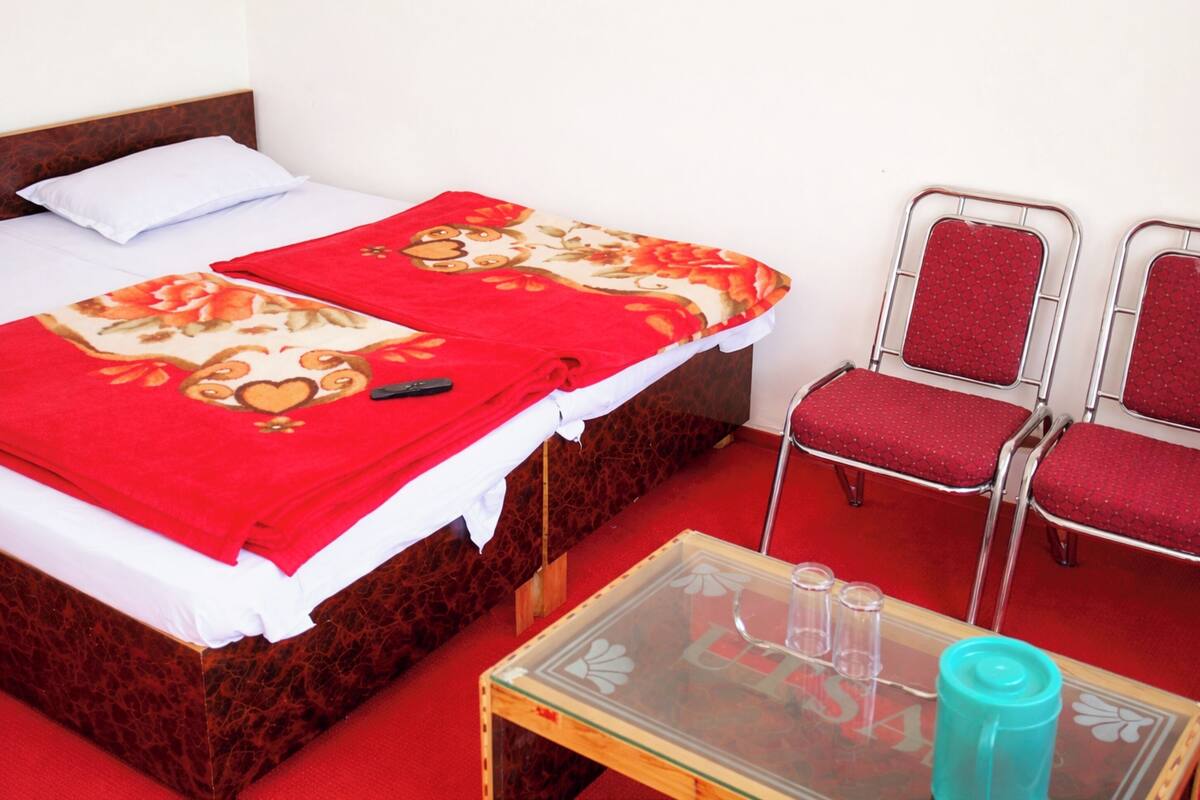 Une chambre d’hôtel avec un lit, deux fauteuils rouges, une petite table avec un plateau en verre et une moquette rouge.