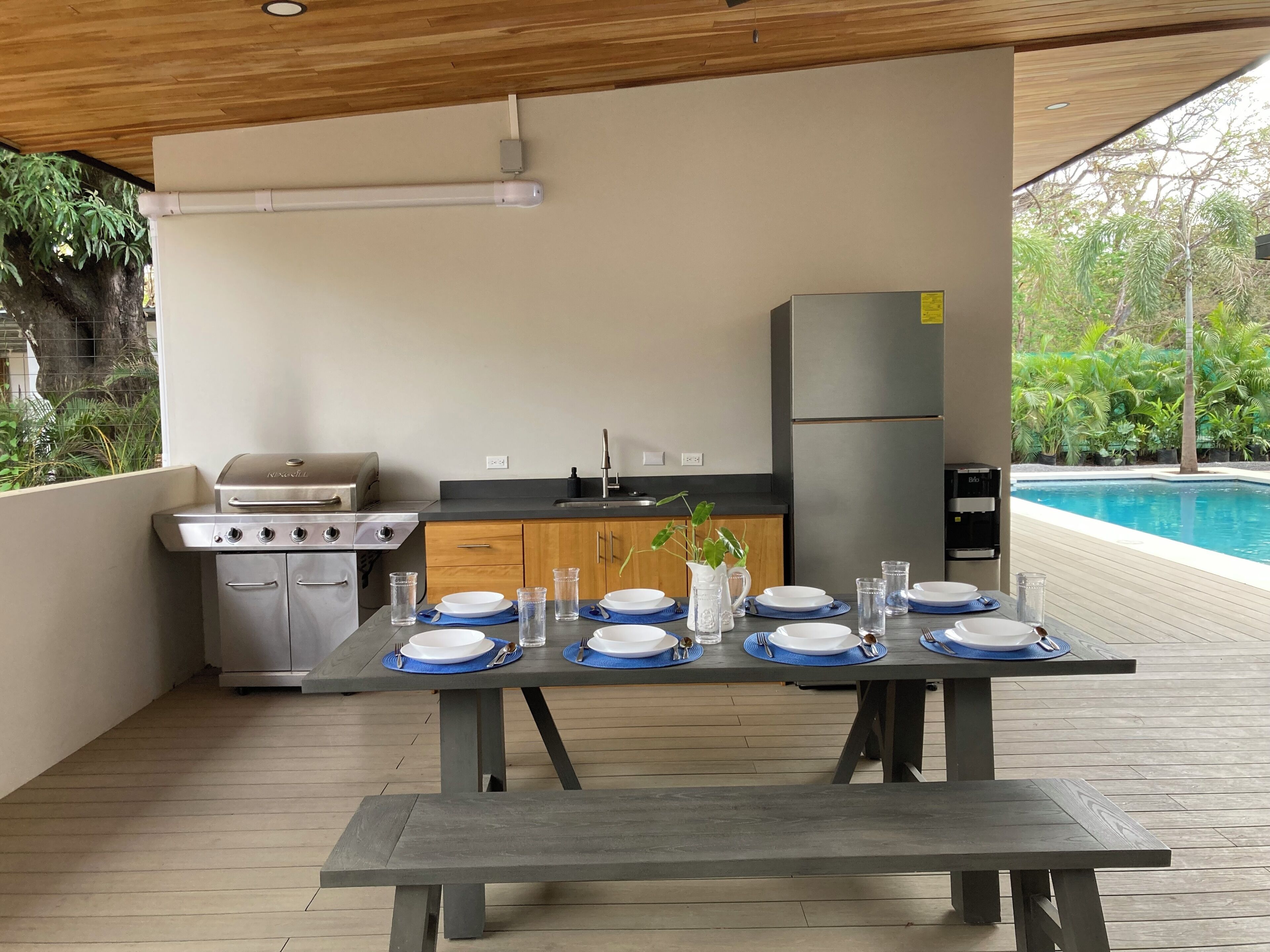 Una cocina exterior con parrilla, pileta y refrigerador, una mesa de comedor dispuesta con platos y vasos, y una piscina al fondo.
