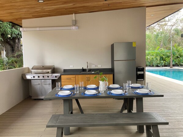 An outdoor kitchen with a grill, sink, and refrigerator, a dining table set with plates and glasses, and a pool in the background. - A Private Sanctuary in Playa Avellanas (Guanacaste Province)