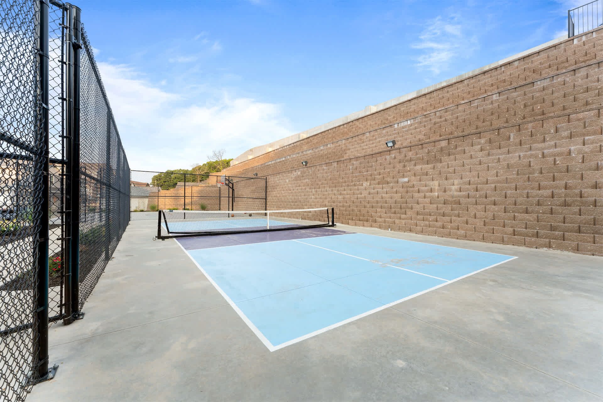 An outdoor tennis court with a chain-link fence, a brick wall, and a clear sky.