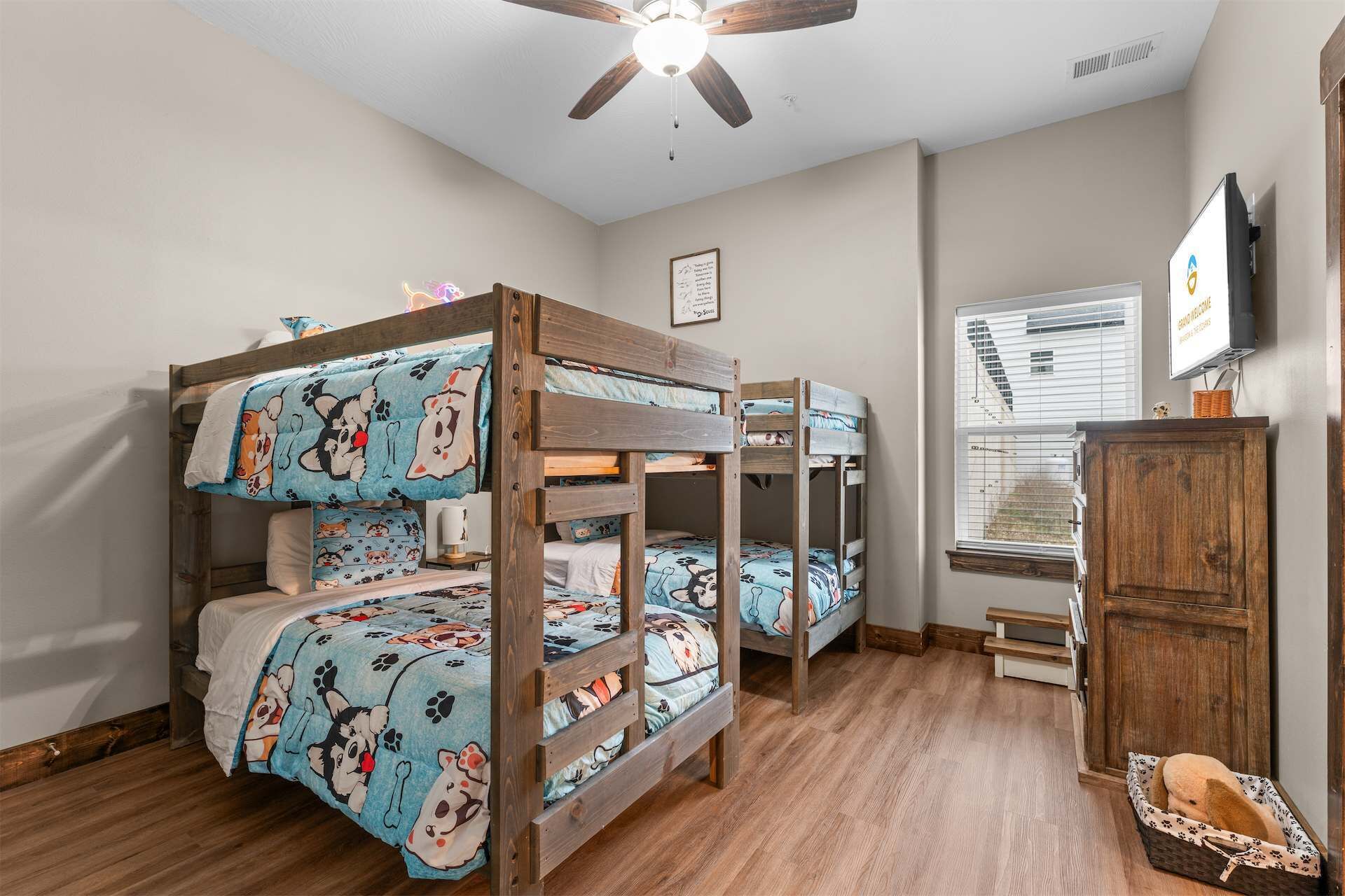 A room with bunk beds, a ceiling fan, a window with blinds, a television, and a wooden cabinet.