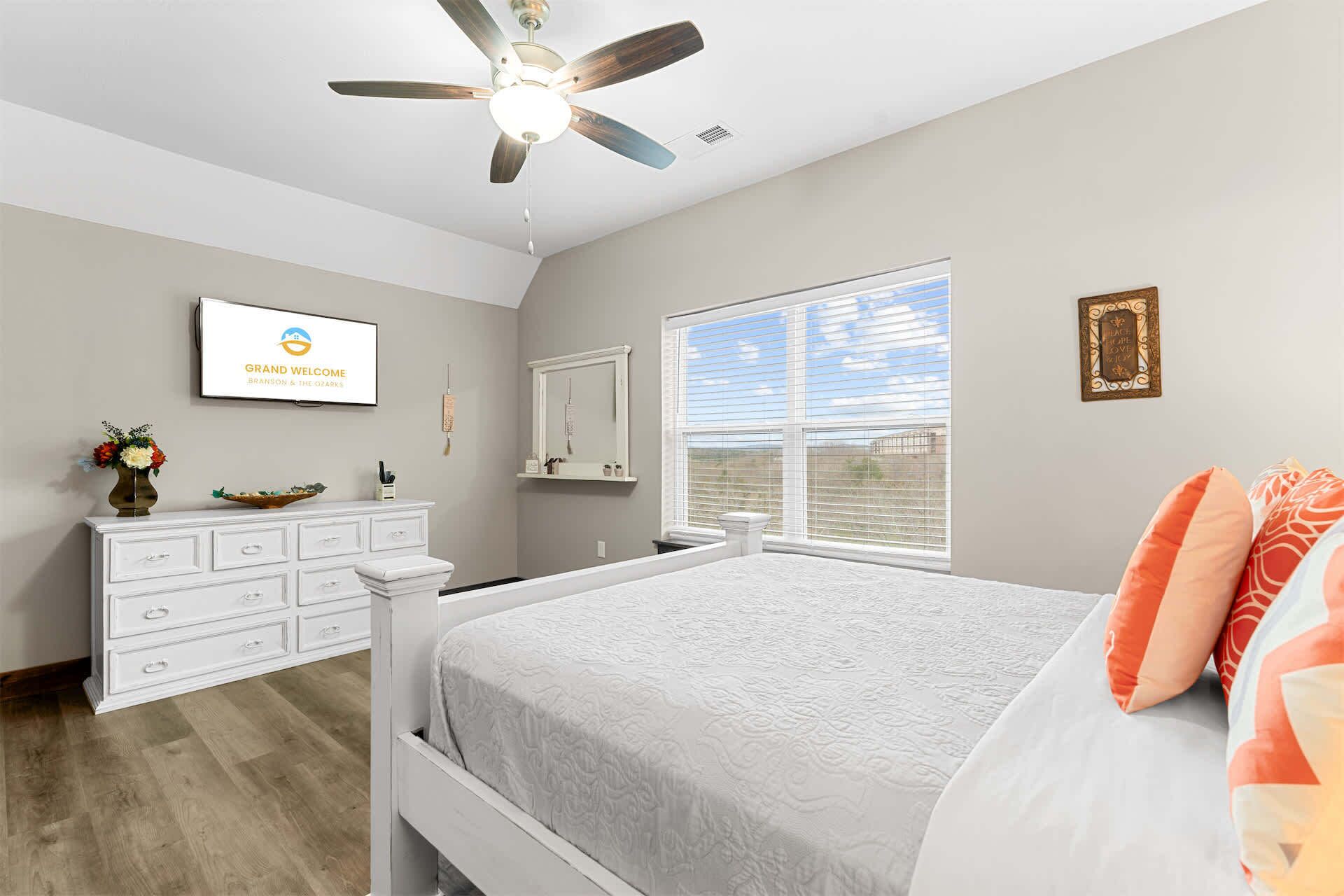 A bedroom with a bed, a ceiling fan, a TV, a window with blinds, a dresser, and a decorative mirror.