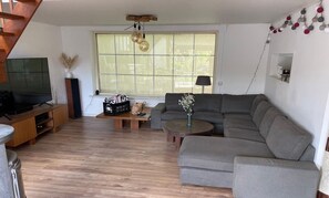 A living room with a grey sofa, a wooden coffee table, a TV on a wooden stand, and a window with a view.