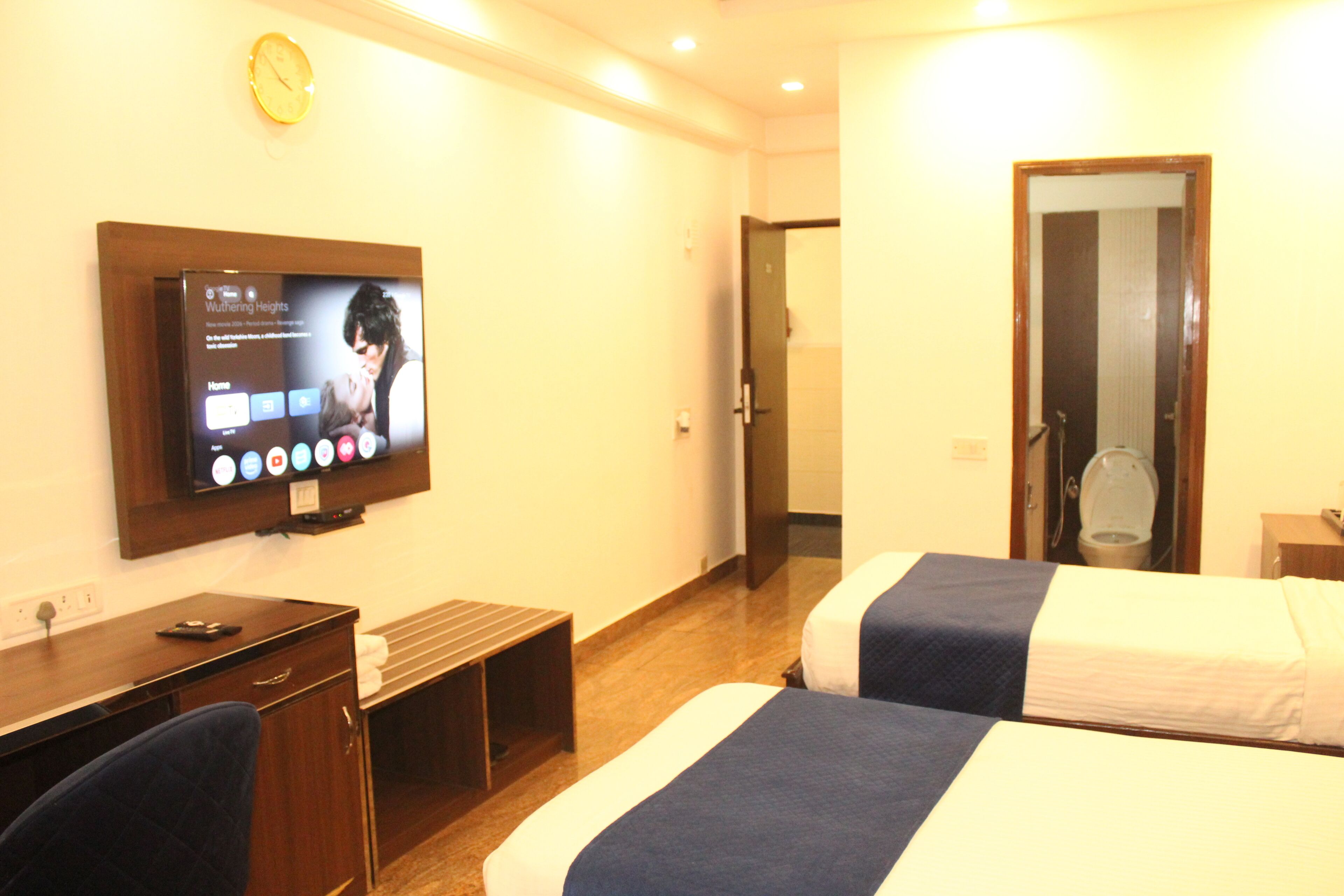 A hotel room with a TV mounted on the wall, a desk, two beds, and a door leading to a bathroom.