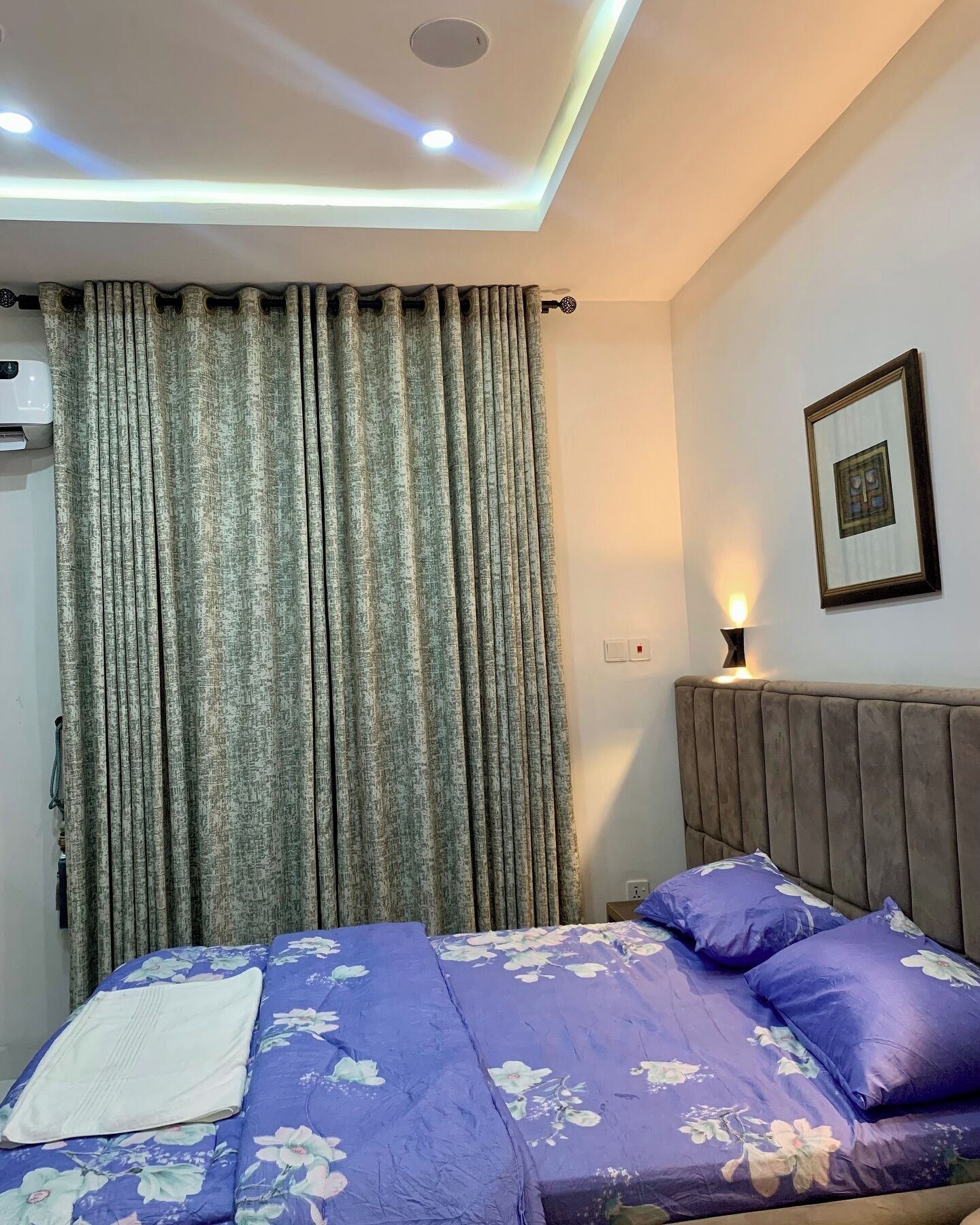 A bedroom with a bed, curtains, a wall-mounted picture, and a bedside lamp.