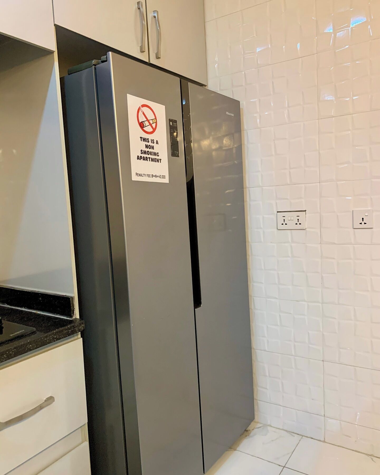 A stainless steel refrigerator with a no-smoking sign in a kitchen with white tiled walls and a black countertop.