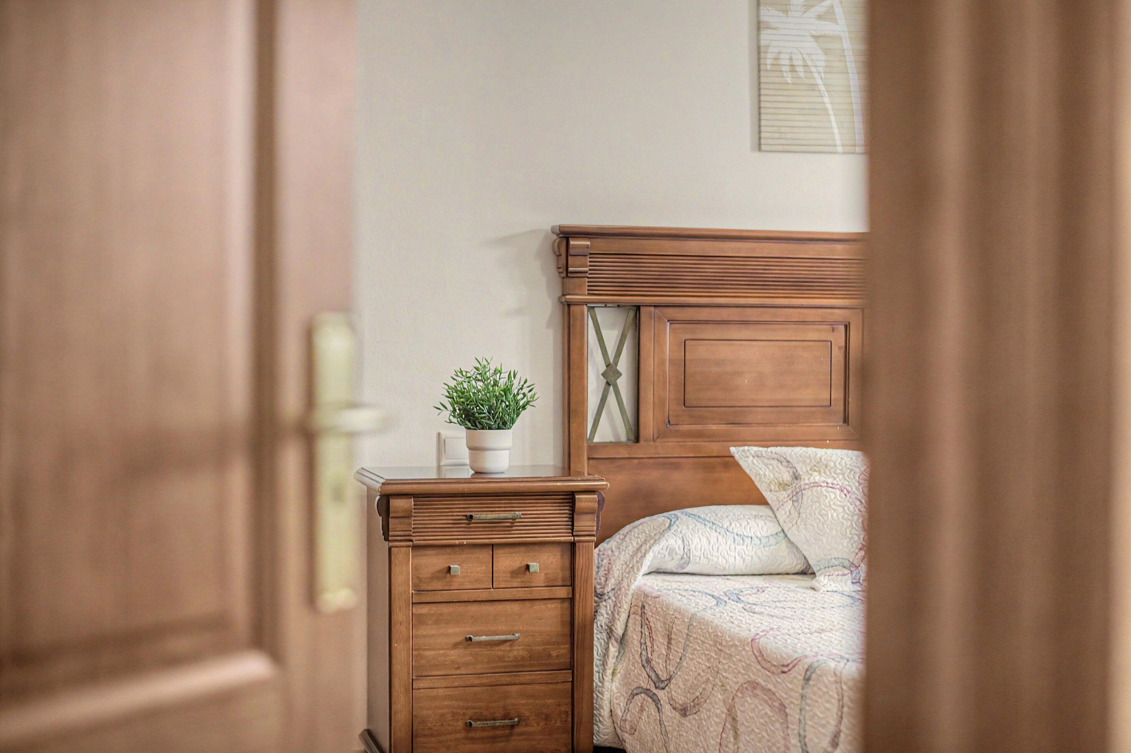 Un dormitorio con cama de madera, una mesita de noche con cajones y una pequeña planta en maceta.