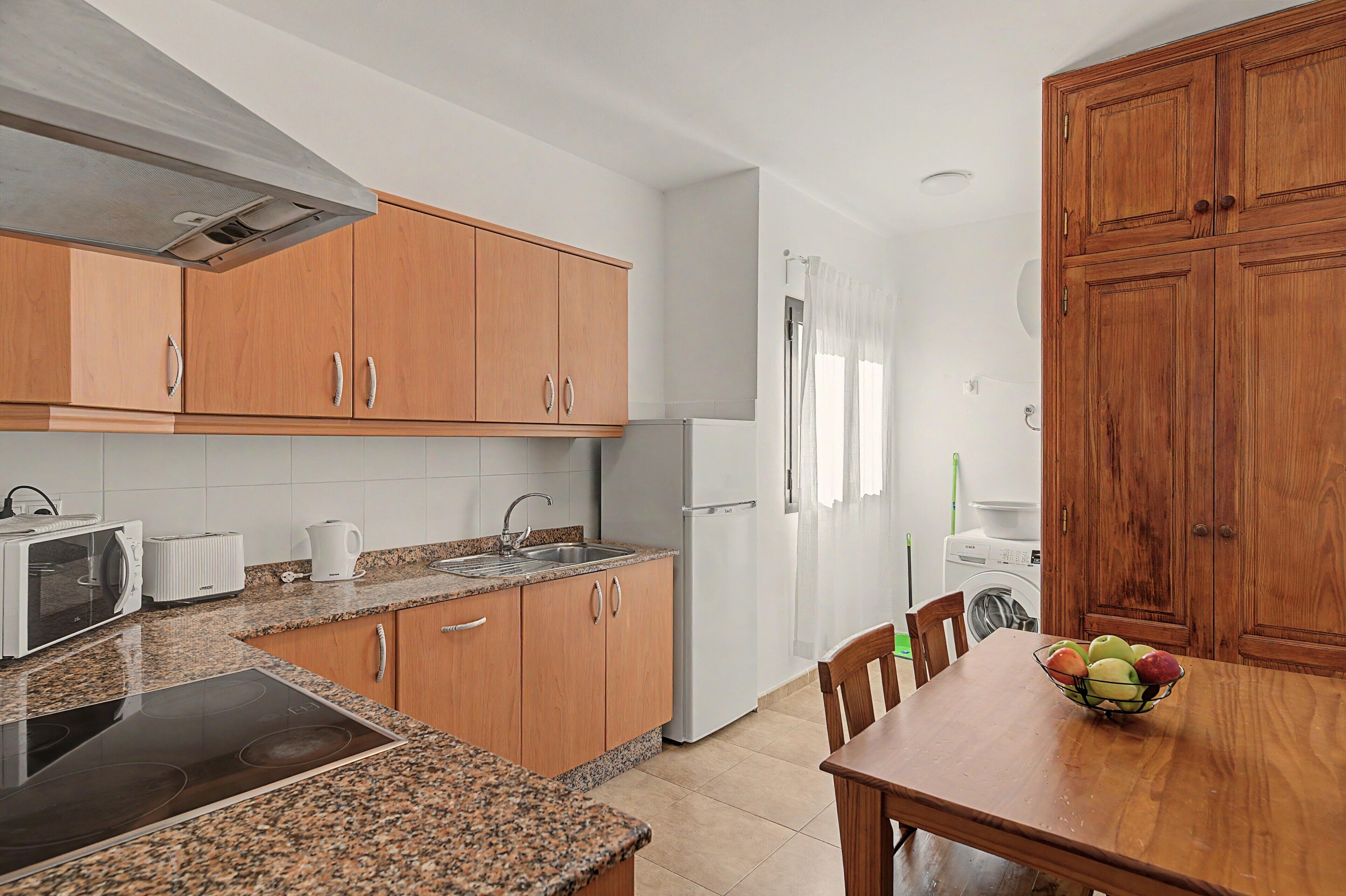 Cocina compacta con armarios de madera, microondas integrado, pileta y comedor con mesa y sillas.