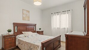 A bedroom with a wooden bed, a nightstand with a plant, a dresser, and a window with curtains.