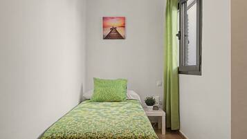 Una habitación pequeña y estrecha con una cama individual, una mesita de noche, una planta en maceta y un cuadro de un atardecer sobre un muelle.