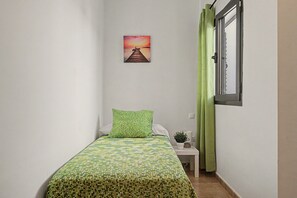 A small, narrow room with a single bed, a nightstand, a potted plant, and a painting of a sunset over a pier.