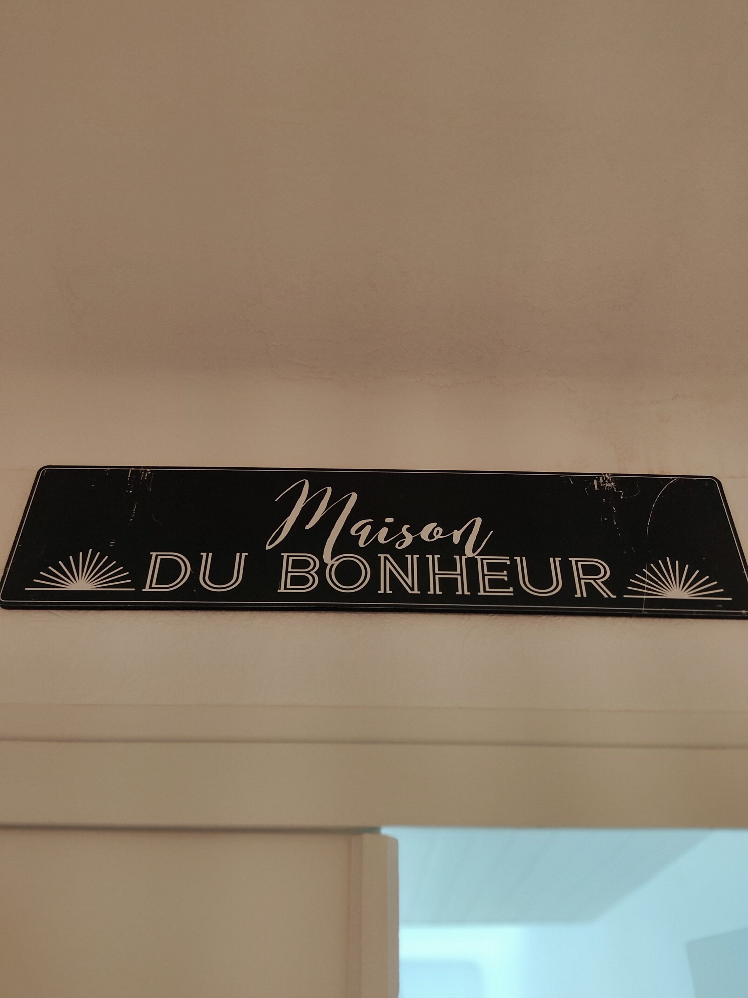 Ein schwarzes Schild mit weißer Schrift, das 'Maison DU BONHEUR' zeigt, mit dekorativen Sonnensymbolen auf beiden Seiten.