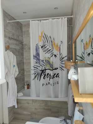 Une salle de bain avec un rideau de douche orné de motifs de feuilles tropicales et de l’inscription « Tropica Paradise », un peignoir blanc posé sur un porte-serviettes et un plancher en bois.