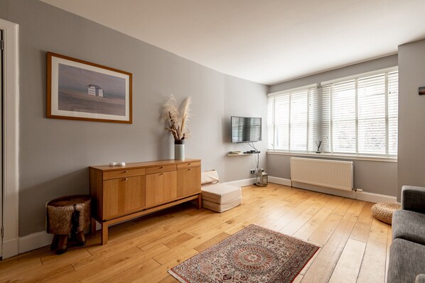 A room with a wooden floor, a sofa, a TV, a radiator, and a window with blinds.