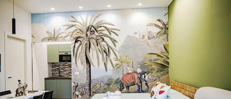 A bedroom with a mural of a palm tree and an elephant, a bed with pillows, a dining table with chairs, and a kitchen in the background.
