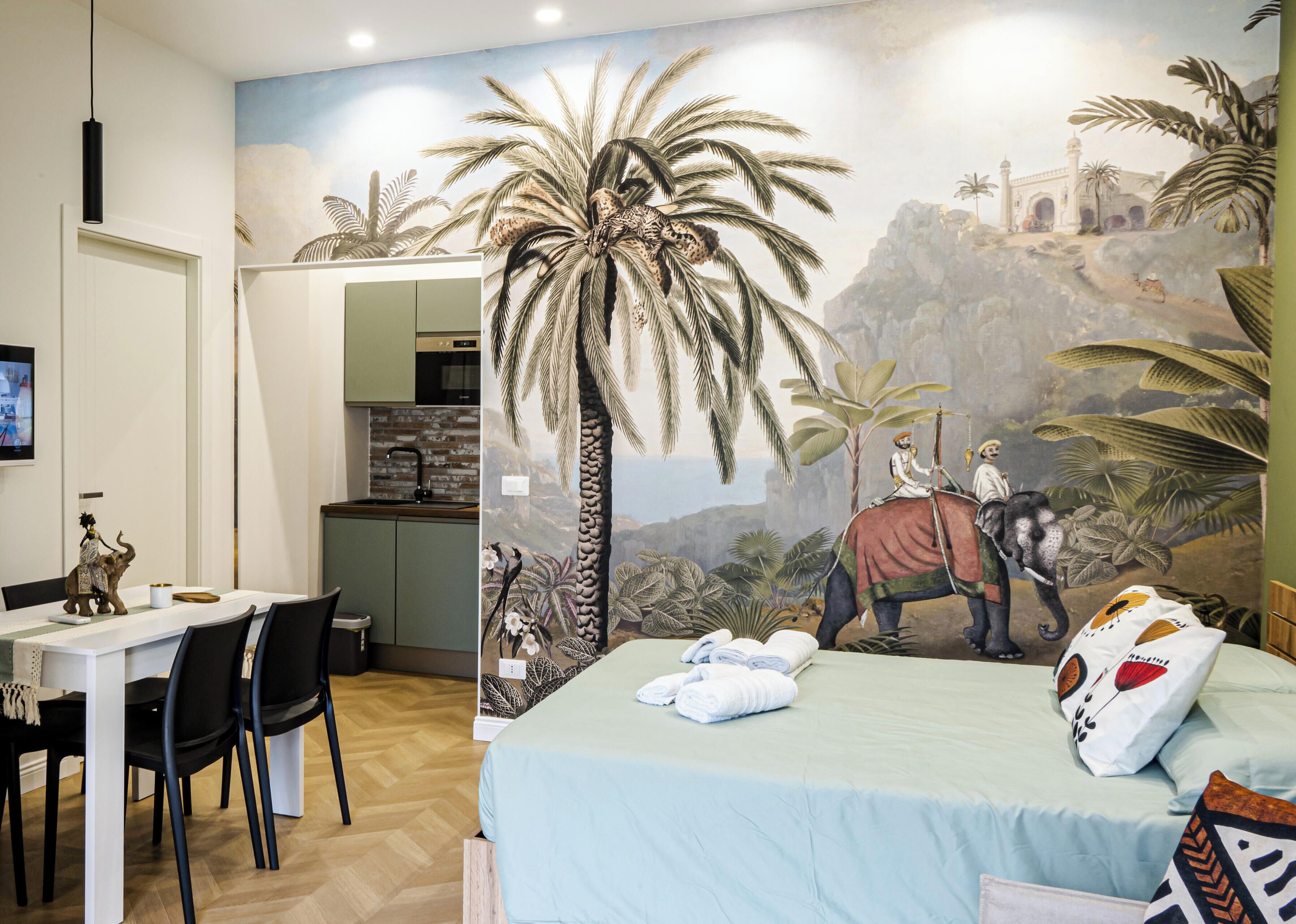 Habitación de hotel con un mural de una escena selvática, una cama con cubrecama azul claro, un escritorio con una silla y una pequeña estatua de elefante.