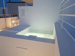Eine Dachterrasse mit einem gläsernen Sitzbereich, einer weißen Wand mit einer Tür und einem Metallgeländer.