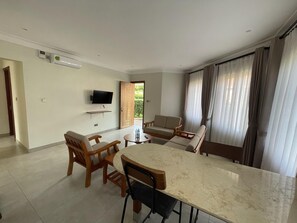Una sala de estar moderna con una mesa de comedor, sillas de madera, un televisor de pantalla plana y un ventanal con cortinas.