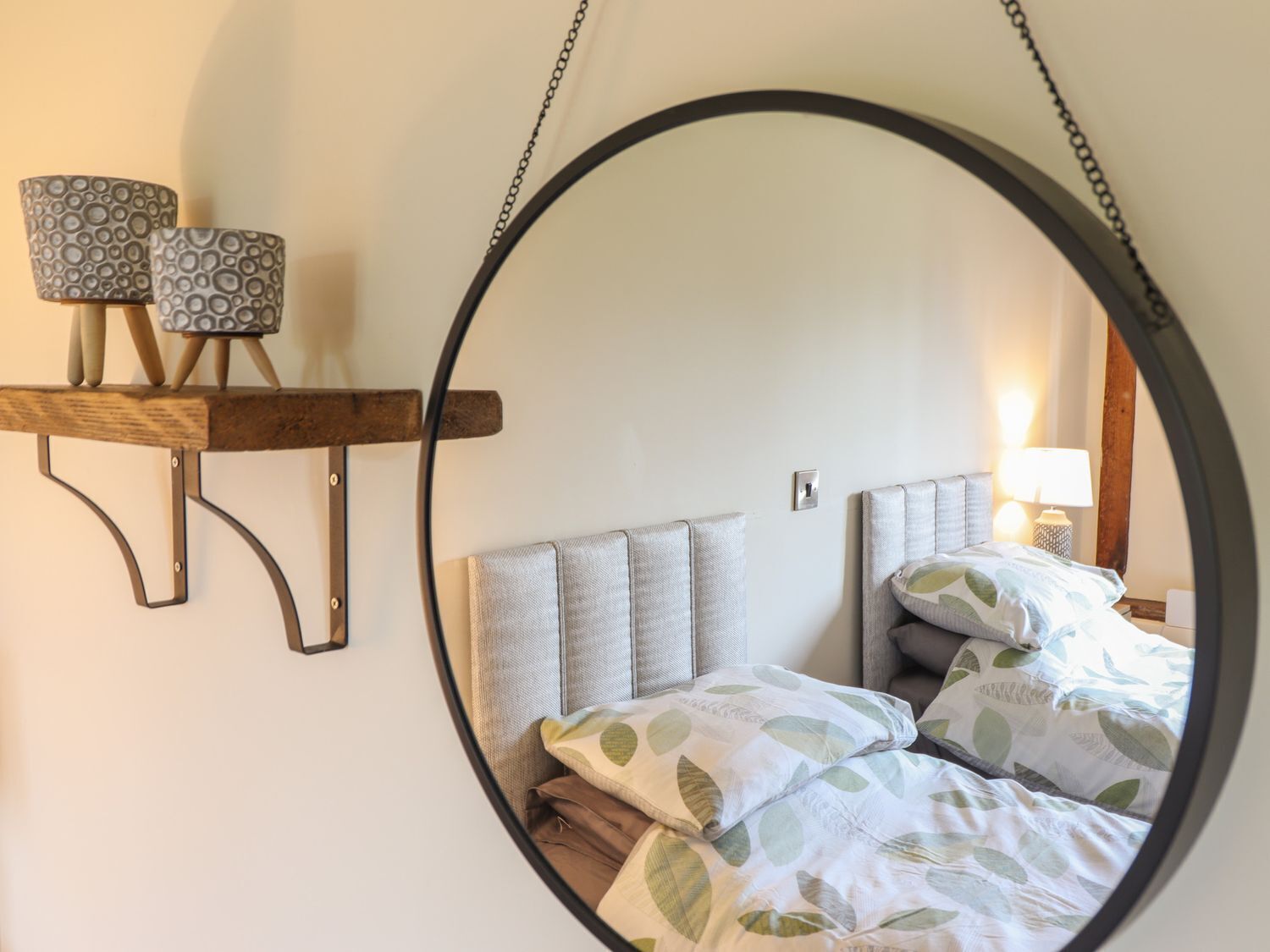 A circular mirror reflecting a bed with leaf-patterned bedding, a bedside lamp, and a shelf with patterned lamps.