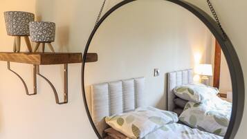 A circular mirror reflecting a bed with leaf-patterned bedding, a bedside lamp, and a shelf with patterned lamps.