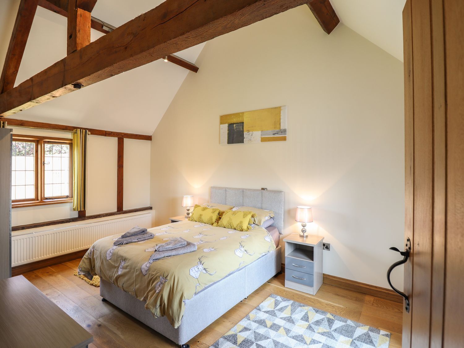 Une chambre à coucher comprenant un lit, des tables de chevet, un radiateur, une fenêtre avec des rideaux et un plafond en pente avec des poutres en bois.