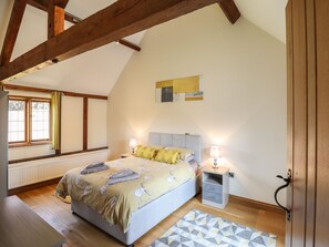 Un dormitorio con cama, mesitas de noche, radiador, ventana con cortinas y techo inclinado con vigas de madera.