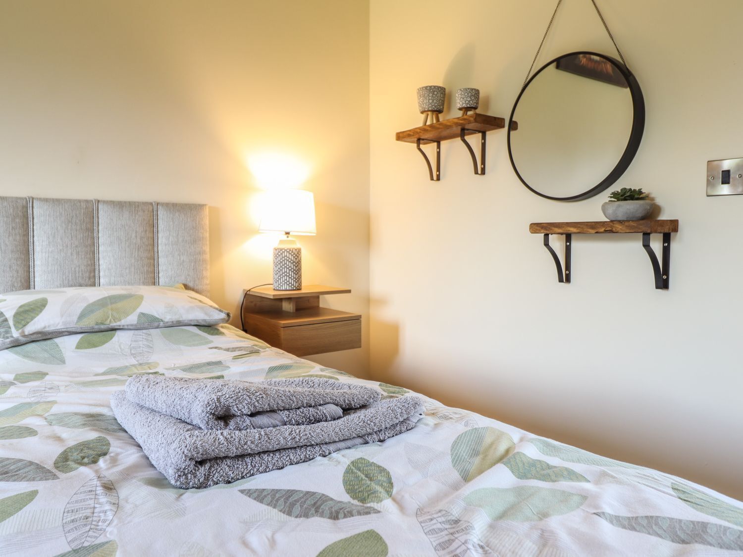 Una cama bien hecha con una colcha de estampado de hojas, una lámpara de noche, un espejo montado en la pared y dos pequeñas repisas con adornos.