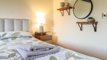Una cama bien hecha con una colcha de estampado de hojas, una lámpara de noche, un espejo montado en la pared y dos pequeñas repisas con adornos.