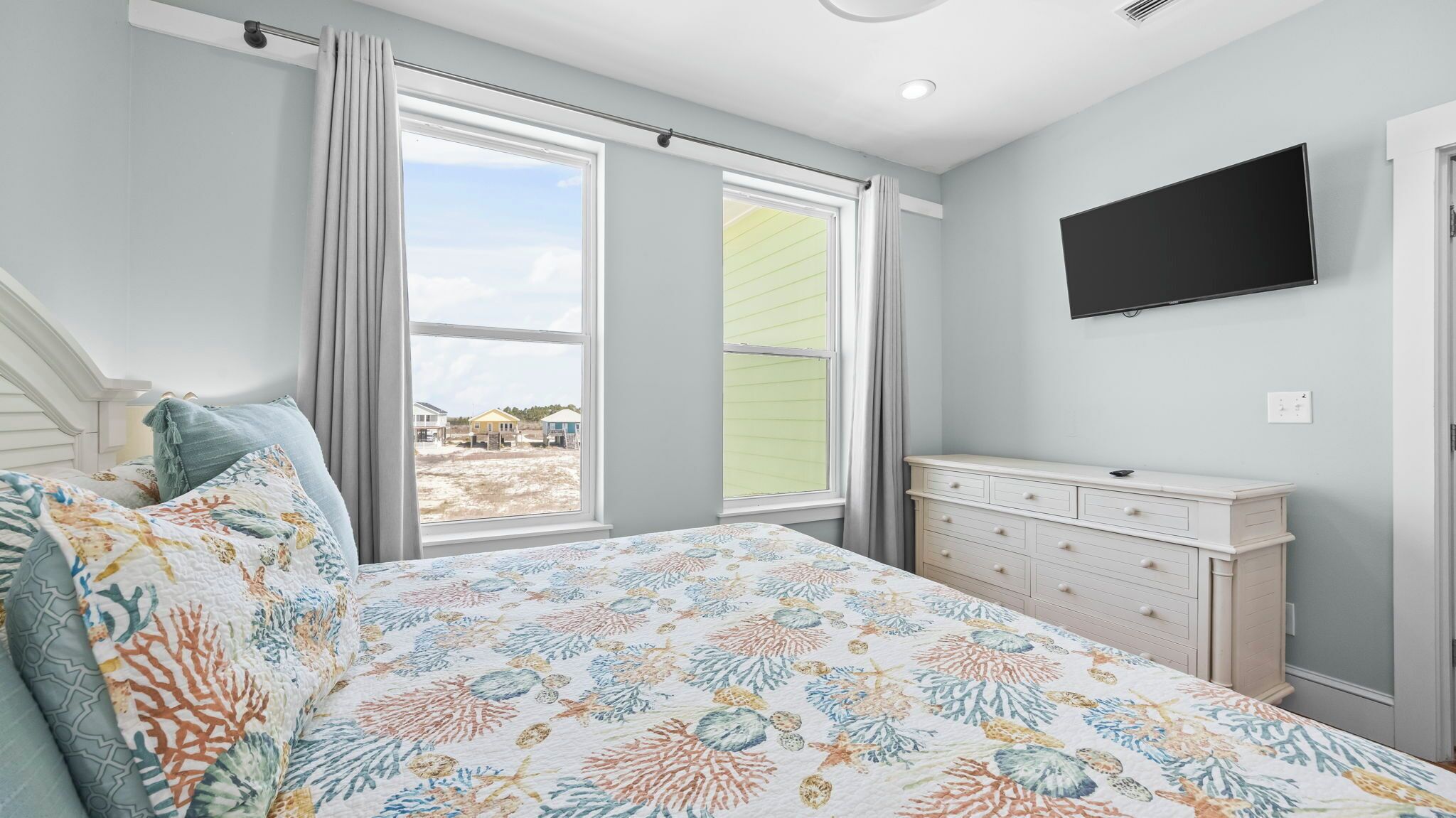 Une chambre à coucher avec un lit, une commode, un téléviseur et une grande fenêtre donnant sur des maisons.