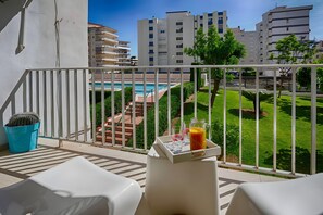 Un balcon avec des balustrades blanches, un cactus, un plateau avec une boisson et une vue sur une piscine et des immeubles.