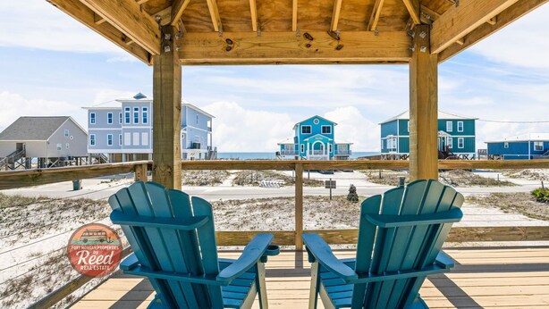 En träveranda med två Adirondack-stolar, med utsikt över en strand där det finns flera hus.